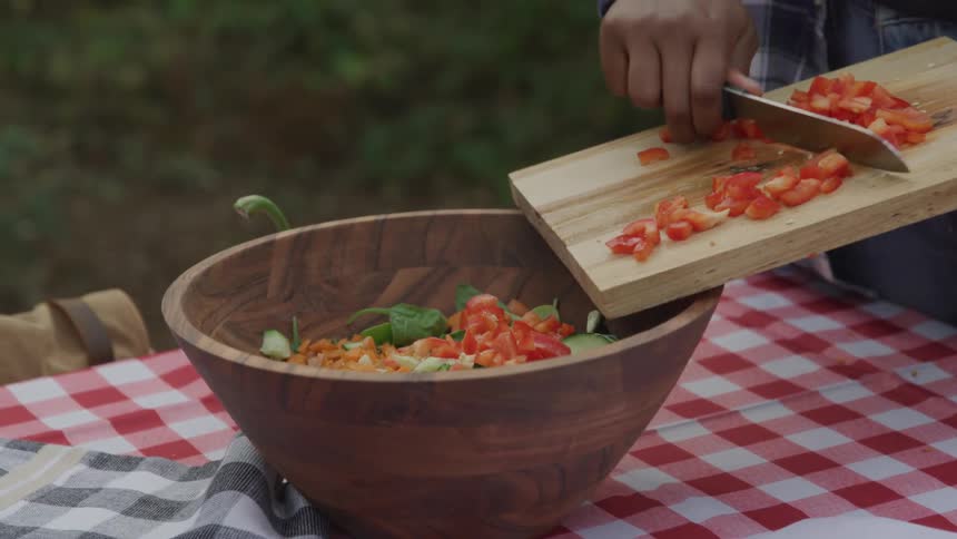 Adding diced tomatoes to a salad sitting on checkered tablecloth outdoors