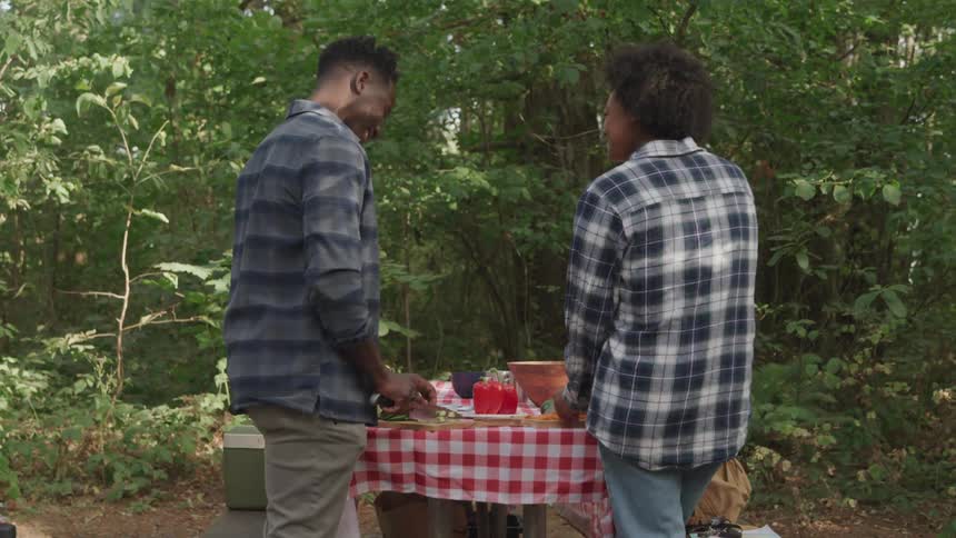 A man and a woman prepare food on a red checkered picnic table amidst lush greenery smiling and enjoying their time together