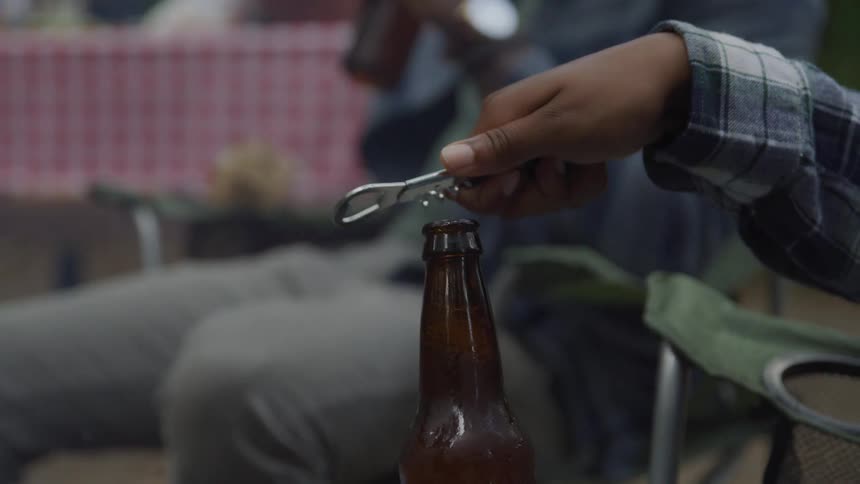 A person in a plaid shirt is using a bottle opener to open a brown beer bottle with another person holding a beer in the background