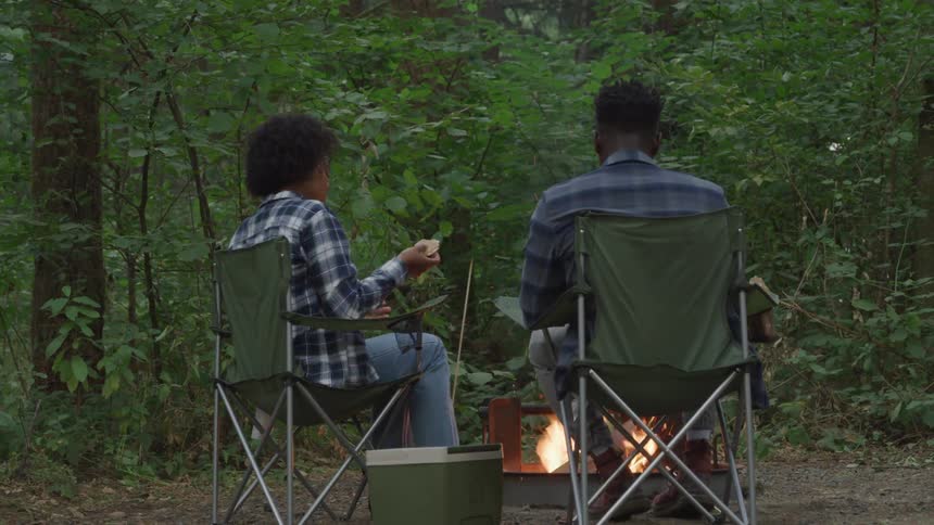 Man and a woman are sitting in camping chairs around a campfire enjoying the natural surroundings of a campsite