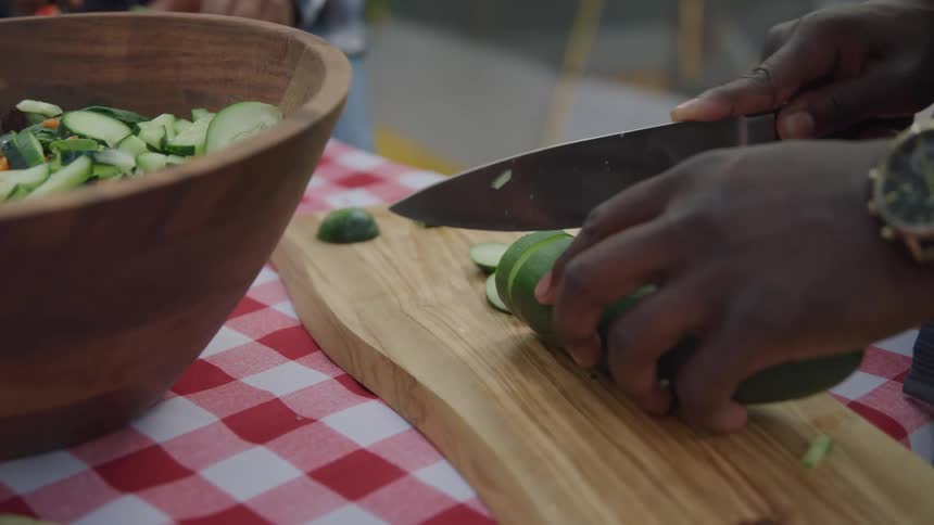 A mans hands slice cucumbers on a wooden cutting board with a wooden bowl filled with salad visible in the background