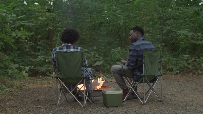 Woman and a man sitting on camping chairs in the middle of nature having lunch with a fire going