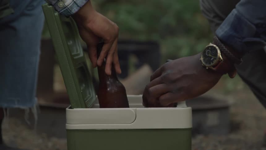 The hands of two men reach into a green cooler outdoors to grab icecold beer bottles with condensation visible on the glass and a fire pit faintly seen in the background