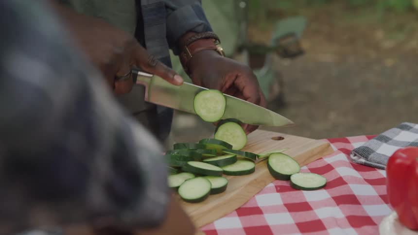 A man miles at woman and slices cucumbers on a wooden cutting board with a red checkered cloth and a red pepper visible in the foreground
