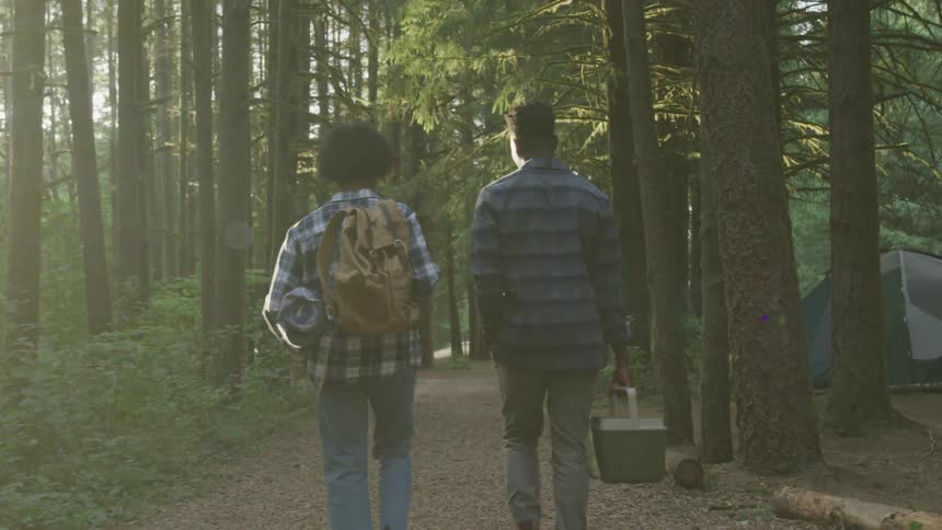 A black couple is walking on a wooded trail while carrying camping gear