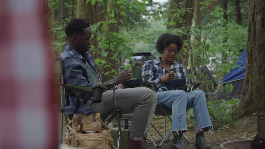 Man and woman stir food in a bowl while sitting at a camp among the trees and talking