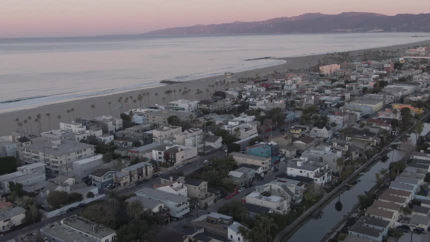 An aerial view of a densely populated urban area adjacent to a sandy beach with the ocean stretching to the horizon and mountains visible in the distance