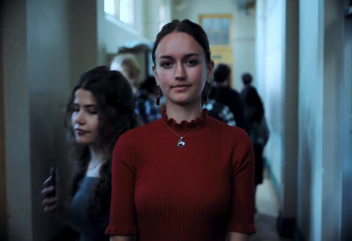 A teenage girl stands still in a school hallway as other kids move past her