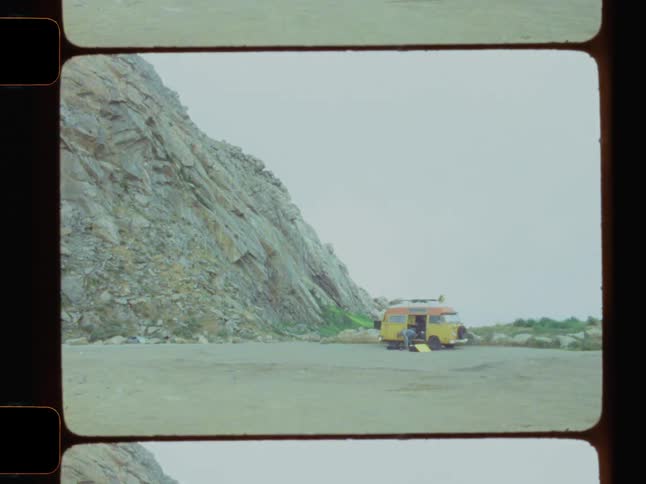 A van is parked on the sandy beach near a cliff under a hazy sky captured on film