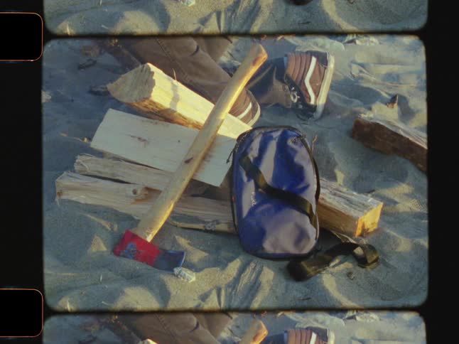 A hand picks a log from a pile of split logs near an axe that lies on the sandy ground and a blue bag