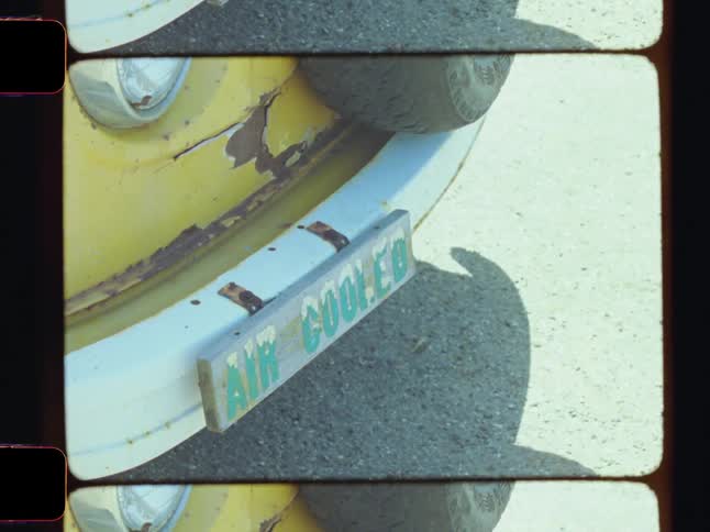 A closeup shows a novelty driver plaque attached to the bumper of an old yellow van parked on a sunny day
