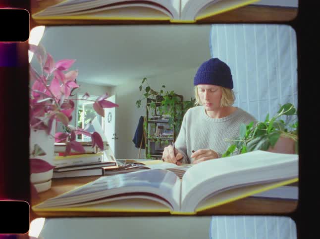 A young man with long blonde hair wearing a blue beanie writes in a notebook sets it on top of a book and begins typing on his laptop