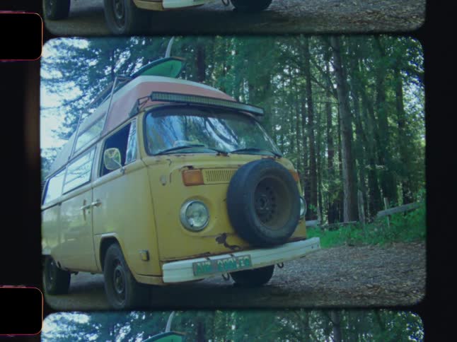 A yellow vintage van with a surfboard strapped to its roof is parked on a dirt path in a forested area
