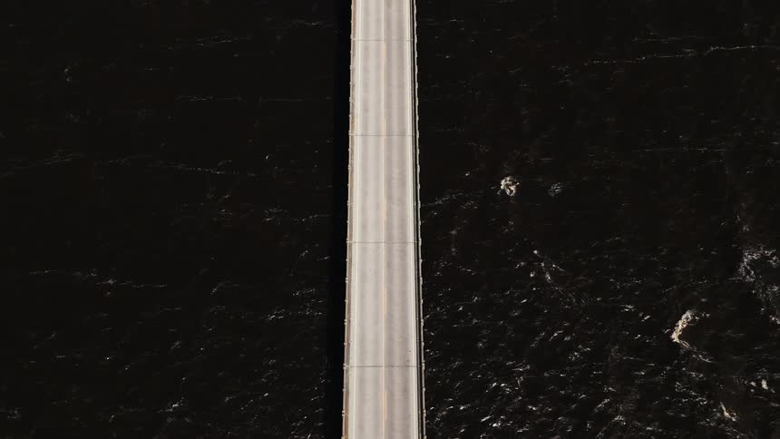 An aerial view shows cars traveling in both directions along a narrow bridge as dark water flows beneath with visible wave patterns and slight surface texture