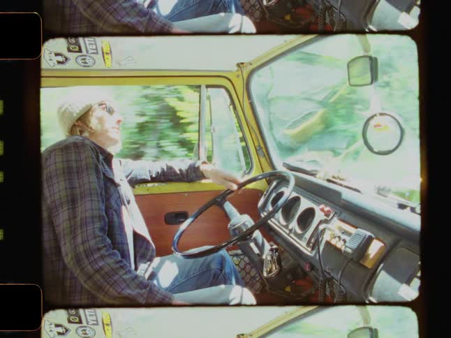 A young man with blonde hair and sunglasses drives a yellow van along a forest on a sunny day