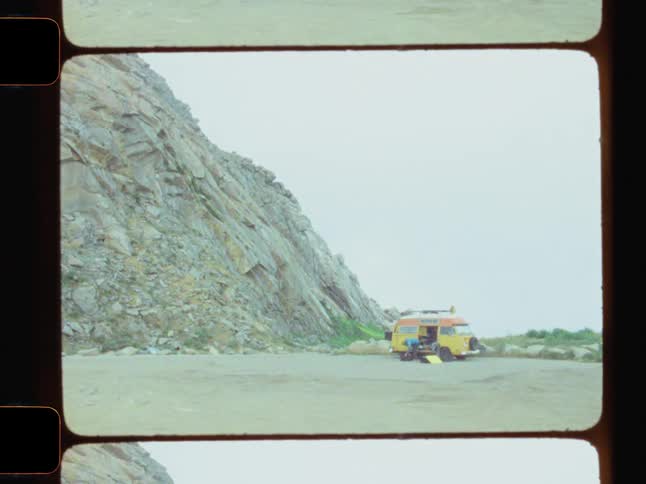 A yellow and orange camper van is parked at the base of a large rocky cliff under an overcast sky