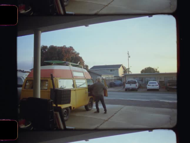 A person stands beside a yellow van with a roof rack closing the drivers door in a residential area with houses and parked cars in the background