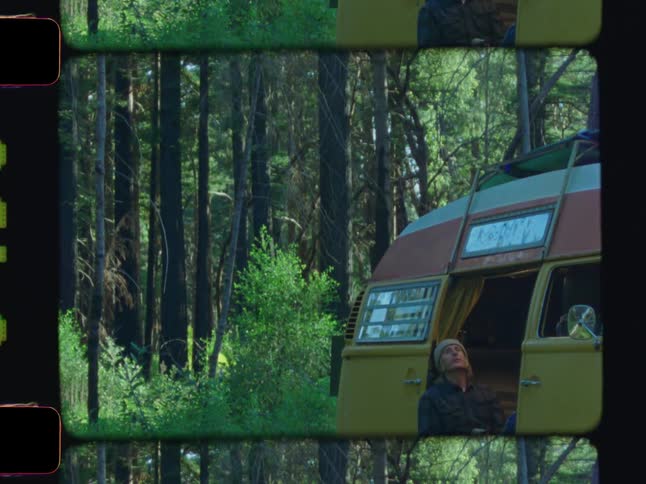 A man sits by the open side door of a vintage van drinking from a cooler in a forest setting
