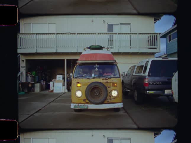 A weathered yellow van with a spare tire on its front bumper is parked on a driveway in front of a lightcolored house with an open garage
