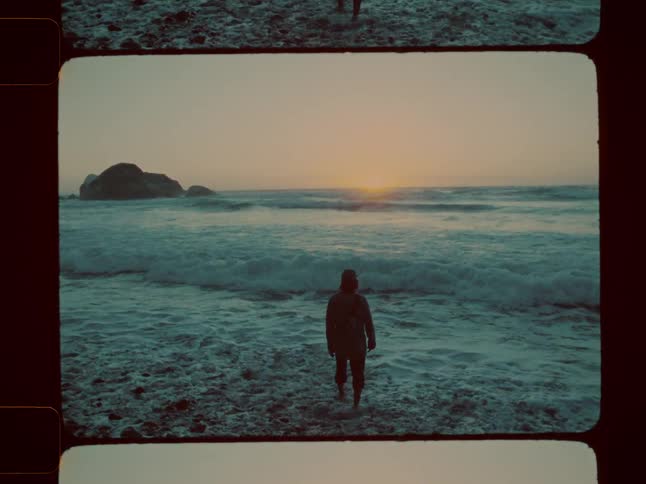 A lone figure stands in the foamy shallows of a turbulent sea at sunset facing the horizon near a rocky outcrop