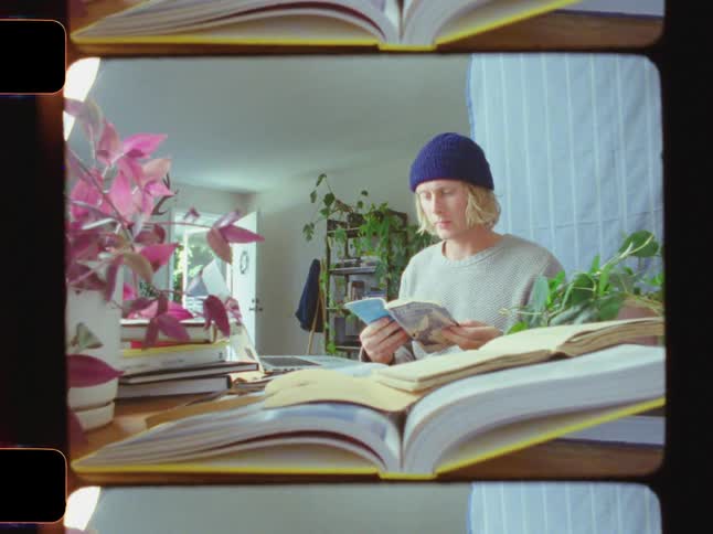 A young man with long blonde hair wearing a blue beanie checks a booklet sets it on top of a book and begins typing on his laptop
