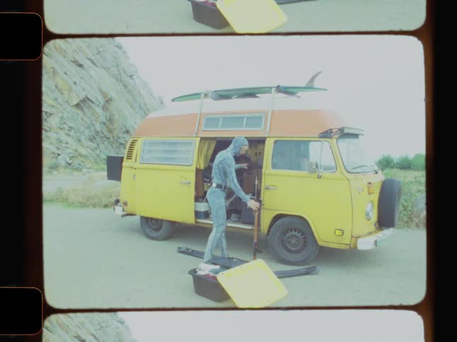 A man in a gray wetsuit gathers underwater fishing equipment from a yellow van parked at the foot of a towering rocky cliff beneath an overcast sky