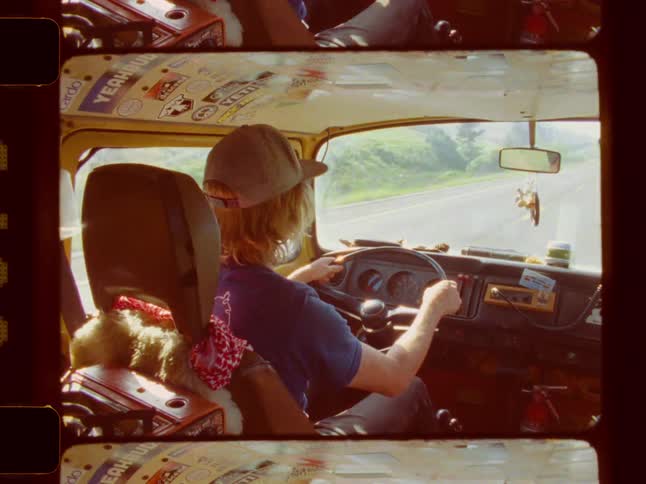 A person wearing a baseball cap drives a stickercovered van on a paved road as seen from the back seat