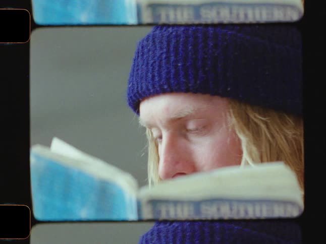 A closeup shows a man with long blonde hair and a blue beanie reading a torn worn book