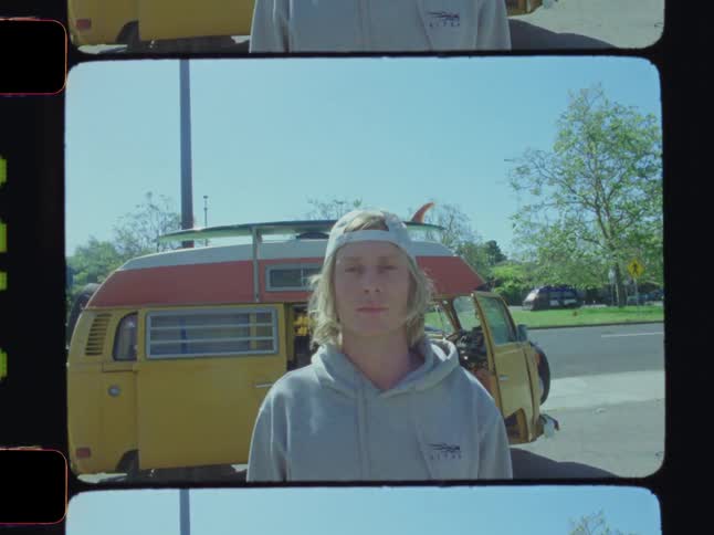 A surfer wearing a grey hoodie is standing in front of his van parked on the side of a street