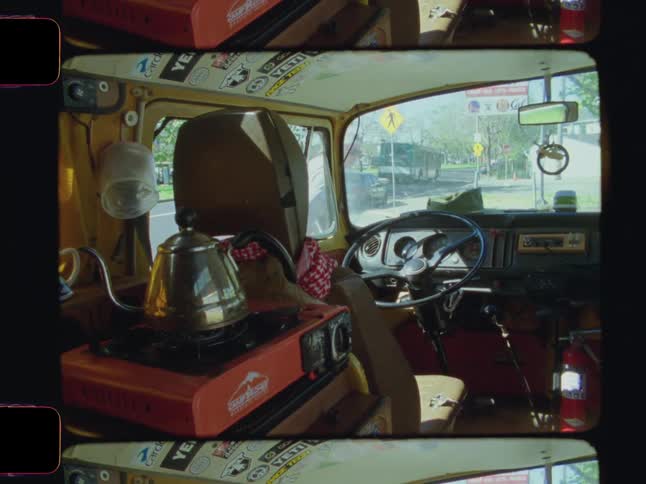 A cramped van interior features a stove teapot stickers and a fire extinguisher while a man steps outside into a sunny day