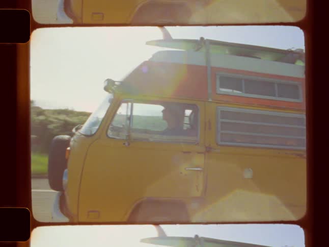 Framed as an analog film shot a vintage yellow volkswagen van with a surfboard on top driven by a white male wearing a beanie is shown