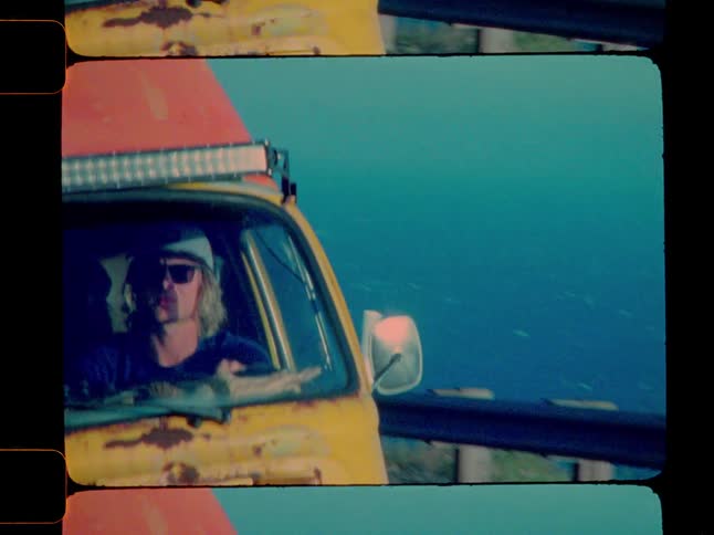 A young man with blonde hair and sunglasses drives a yellow van along the oceans edge