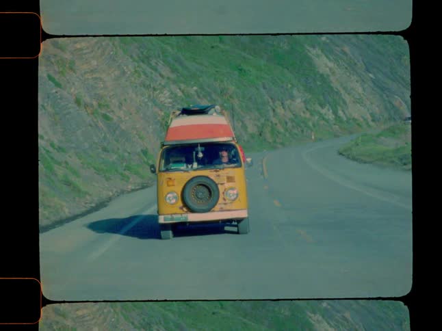 A vintage yellow volkswagen camper van with a red and white striped top travels along a winding paved road with a backdrop of a hillside area seen off in the distance