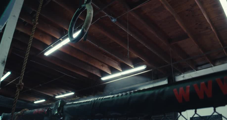 Gymnastic rings hang from the ceiling beams above a section of a fighting cage in a dimly lit training facility