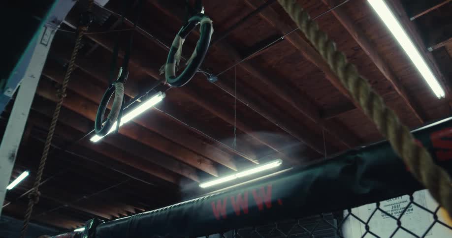 Gymnastic rings hang from the ceiling beams above a section of a fighting cage in a dimly lit training facility