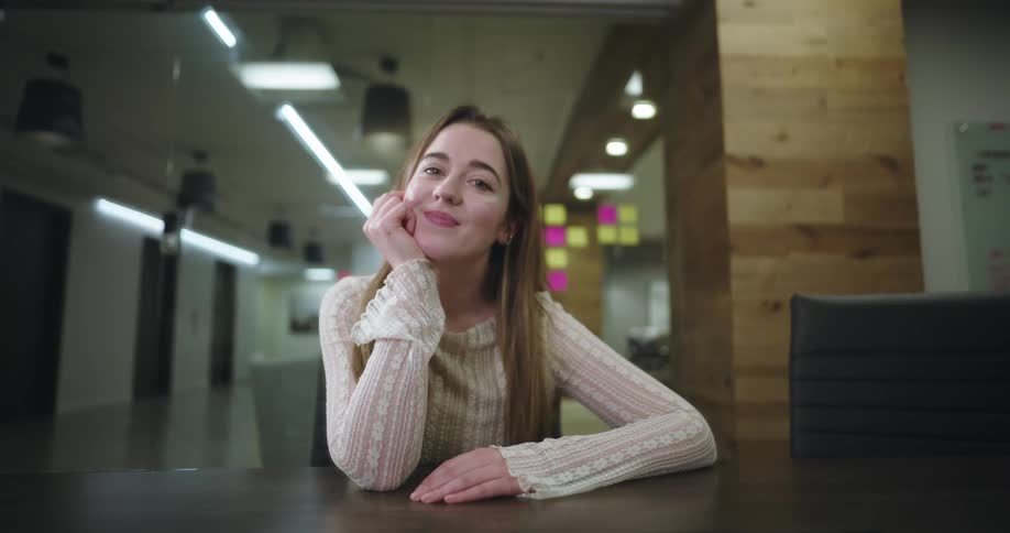 A blondehaired woman smiles while seated at a table in a modern office conference room looking towards the viewer against a background with a glass wall