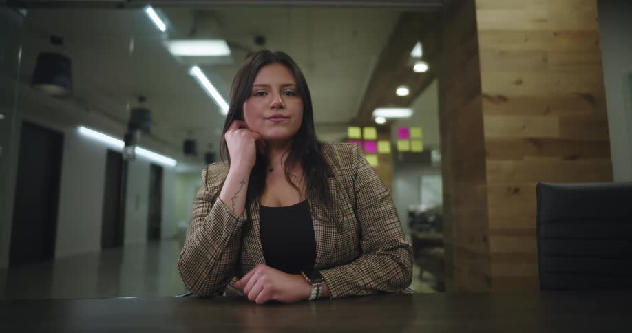 A woman with long hair is seated in a welllit modern waiting area looking at the camera with background elements such as a glass wall out of focus