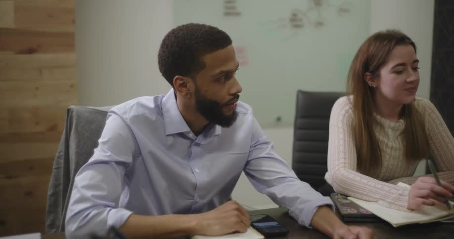 A group of diverse professionals are engaged in a meeting around a table with a man in a blue shirt as the primary focus as the camera focuses on him and his reaction to the meeting