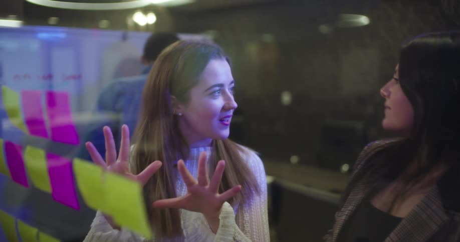 A person stands in front of a glass wall covered with colorful sticky notes gesturing animatedly while another person observes nearby