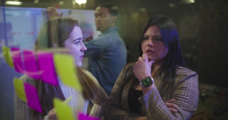 The two woman are looking at post it notes and talking to each other and behind them is a male who is listening to them and writing on a white board