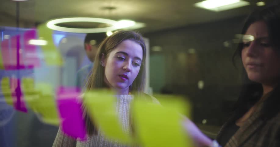 Two women are engaged in a conversation in a modern office environment positioned behind a glass wall with colorful elements
