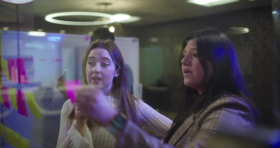 Two women are engaged in a discussion in a modern office setting standing before a glass board likely containing notes