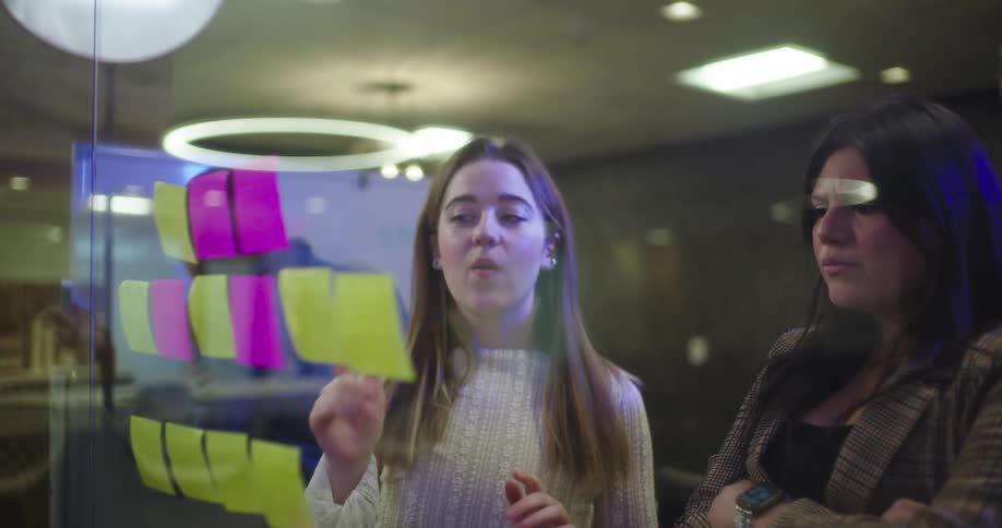 Two women talk as they point to notes hanging on a window in an office