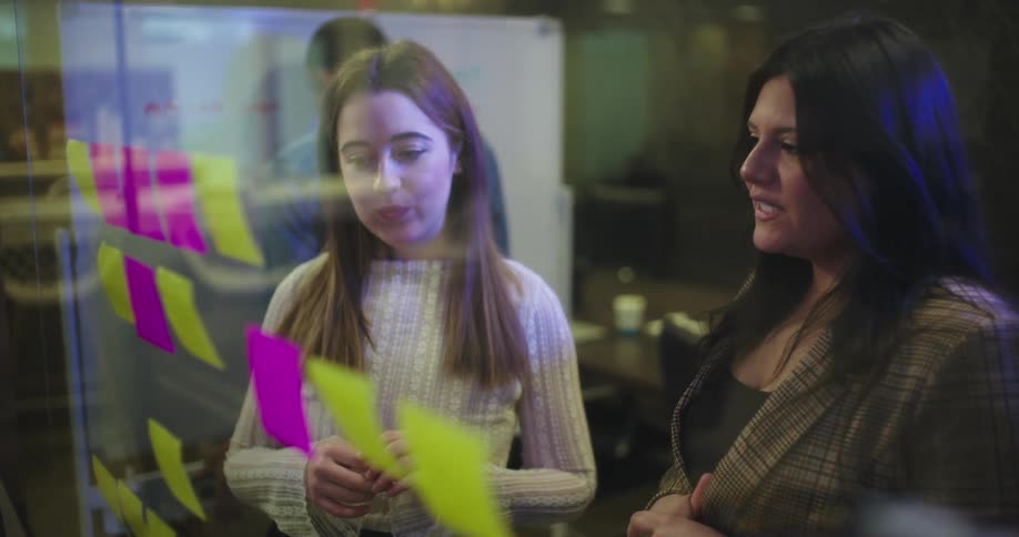 Two women work with sticky notes on a clear board in an office