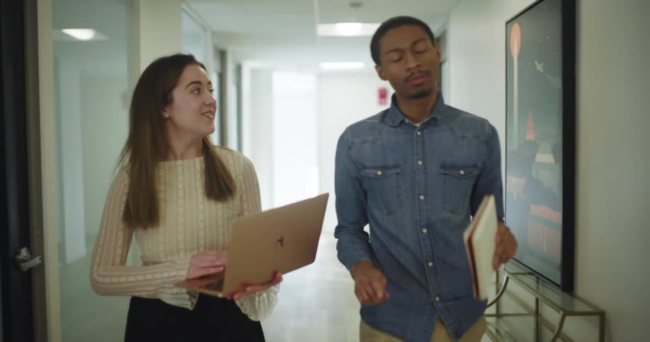 A woman holding a laptop and a man carrying a notebook walk together down a hallway engaged in conversation