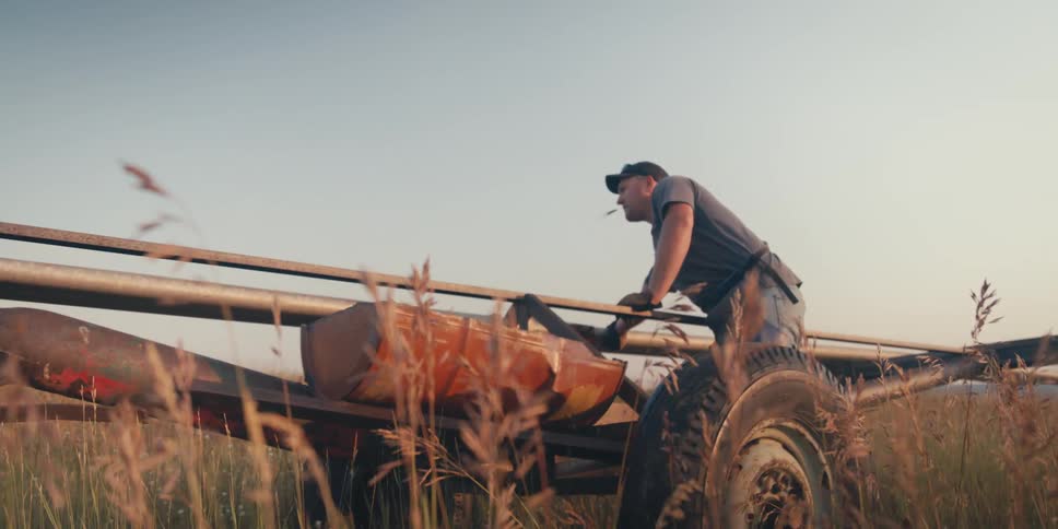 The man in the hat is using a type of farm equipment in a hay field and another type of equipment drives behind him then out of sight again