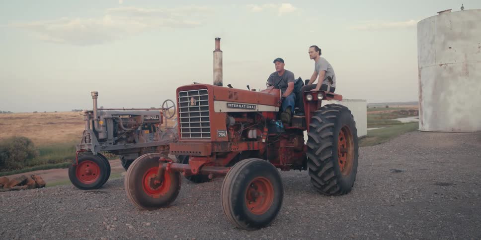 Bathed in the warm light of sunset two farmers sit upon their vintage tractors in a rural setting seemingly engaged in a relaxed conversation