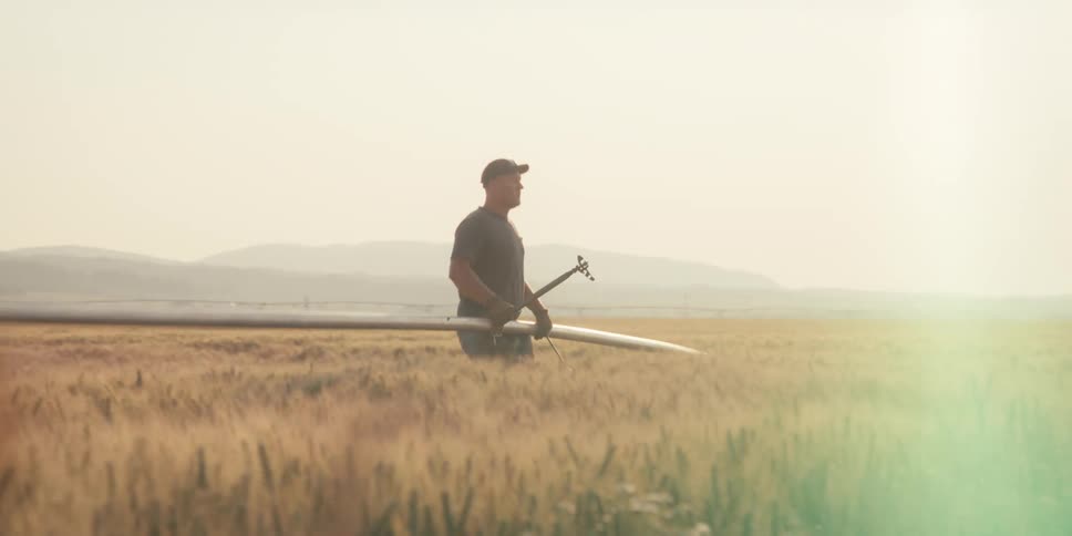 A farmer walks through a wheat field at sunset carrying a long irrigation pipe with the pipes edge bouncing slightly as he moves toward a tractor under a hazy sky and distant rolling hills