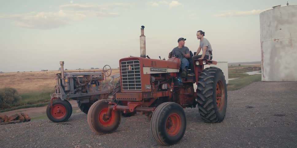 Two men sit on a farming tractor next to a grassy field