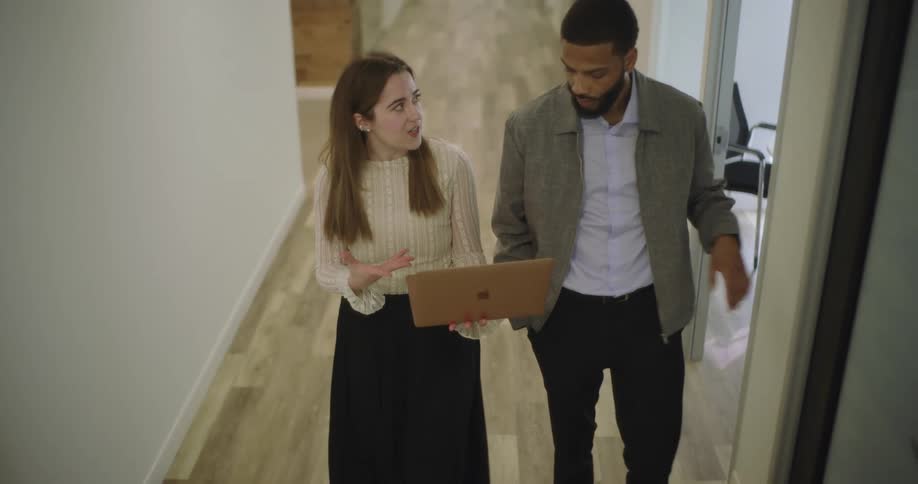 A man and woman talk and look at a laptop as they walk down the hallway in an office